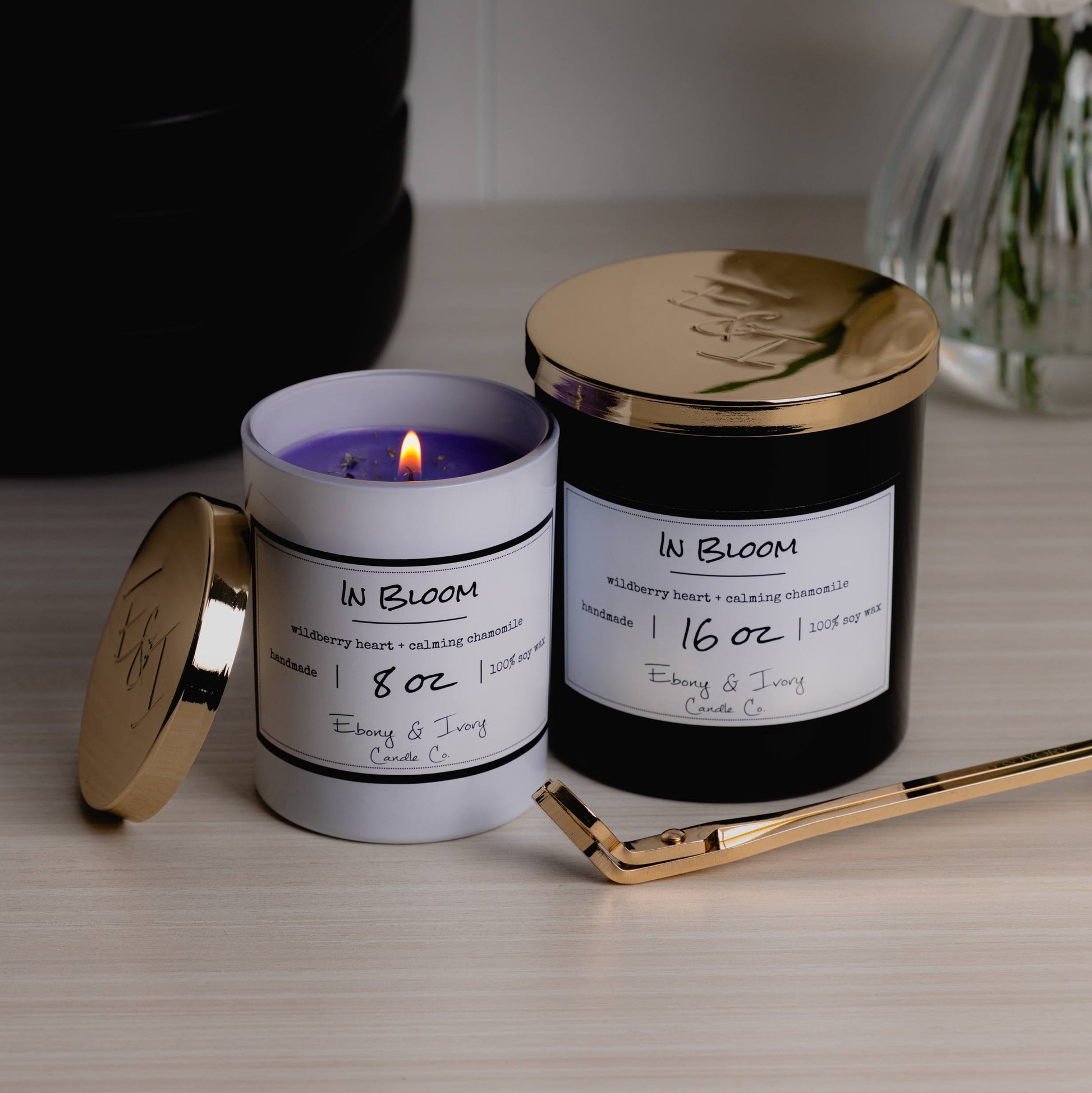 Two candles sitting next to eachother. The first one is a white 8oz jar soy candle with a gold lid sitting next to it, the wick is lit and it show cases purple coloured wax with wildflowers on top, it has a white label named In Bloom. The second candle is a black 16oz jar soy candle with a gold lid on top and a white label named In Bloom, they're sitting on a wood surface with gold wick trimmers nearby and a vase of flowers behind. They're made by Ebony & Ivory Candle Co in Lethbridge, Alberta, Canada