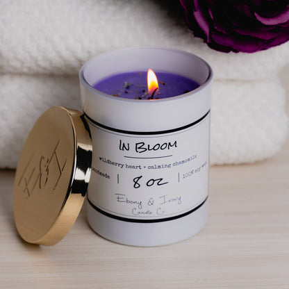 A white 8oz jar soy candle with a gold lid and a white label named In Bloom, on a wood table with fluffy white towels and purple flowers behind it, it had purple wax and mixed dried flower petals on top, made by Ebony & Ivory Candle Co in Lethbridge, Alberta, Canada