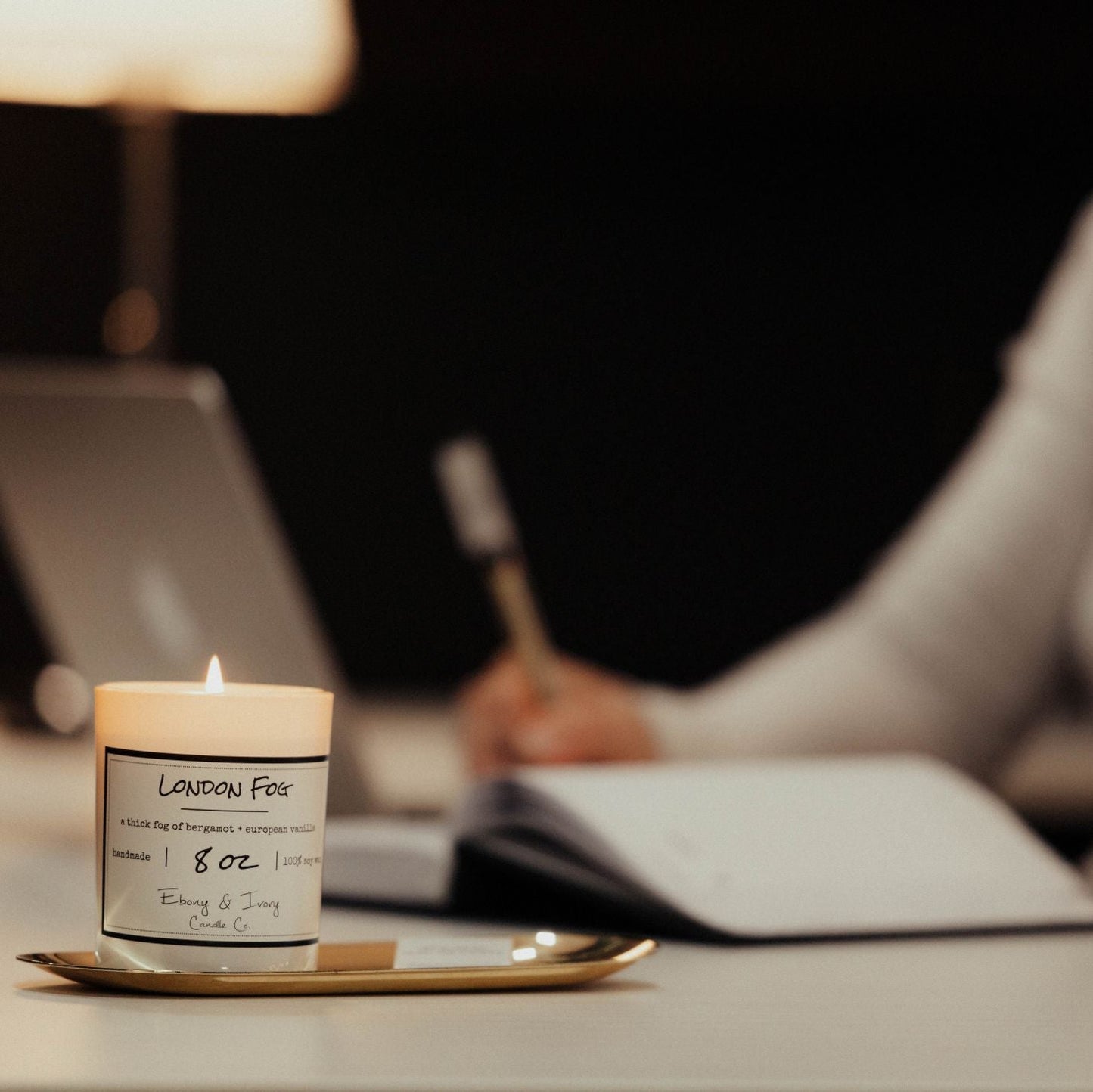 A white jar 8oz soy candle with a gold lid and a white label named London Fog, on a white office desk with a person working on a laptop and a notebook behind it, made by Ebony & Ivory Candle Co in Lethbridge, Alberta, Canada