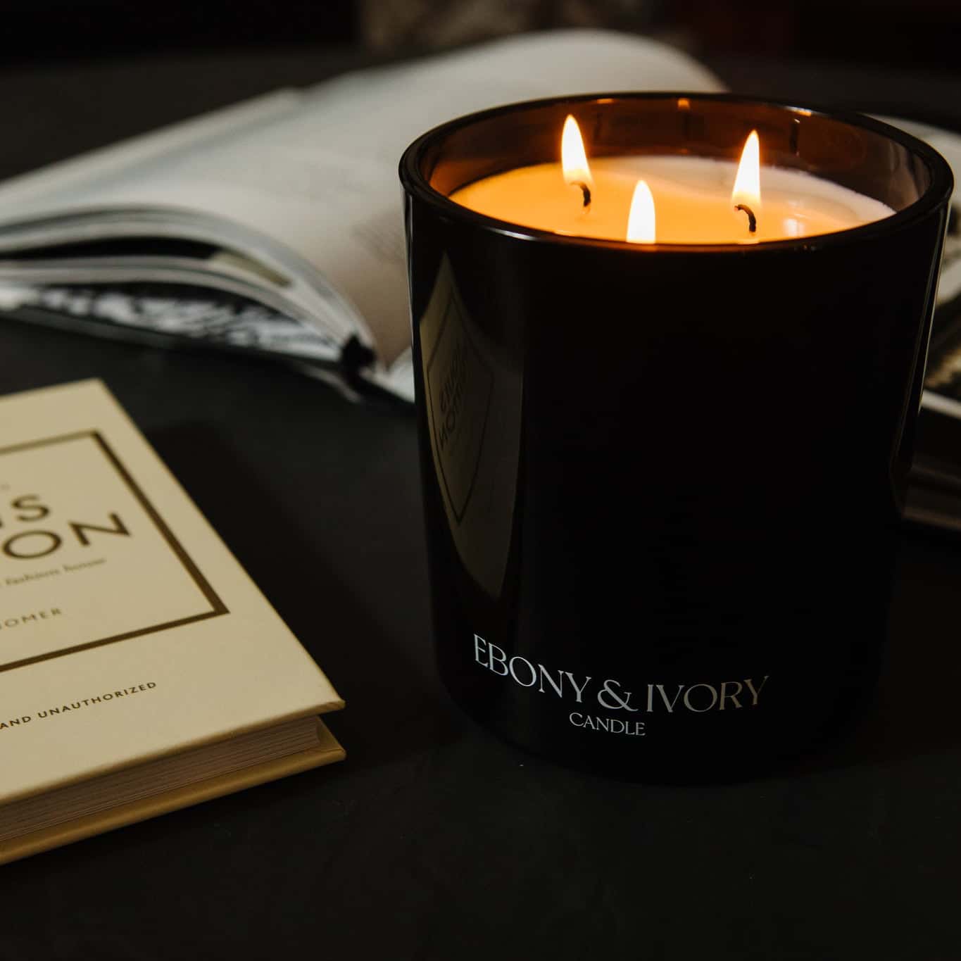 A black 18oz jar, 3 cotton wick, coconut soy candle with a white Ebony & Ivory on the front, on a black table with open books around it, made by Ebony & Ivory Candle Co in Lethbridge, Alberta, Canada