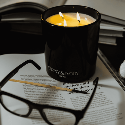 A black 18oz jar, 3 cotton wick, coconut soy candle with a white Ebony & Ivory on the front, on an open book with black rimmed glasses, with open books around it, made by Ebony & Ivory Candle Co in Lethbridge, Alberta, Canada