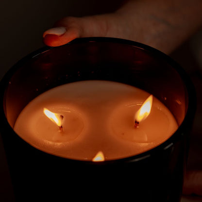 A black 18oz coconut soy candle with three lit wicks being held by a woman with white nails. Made in lethbridge alberta canada
