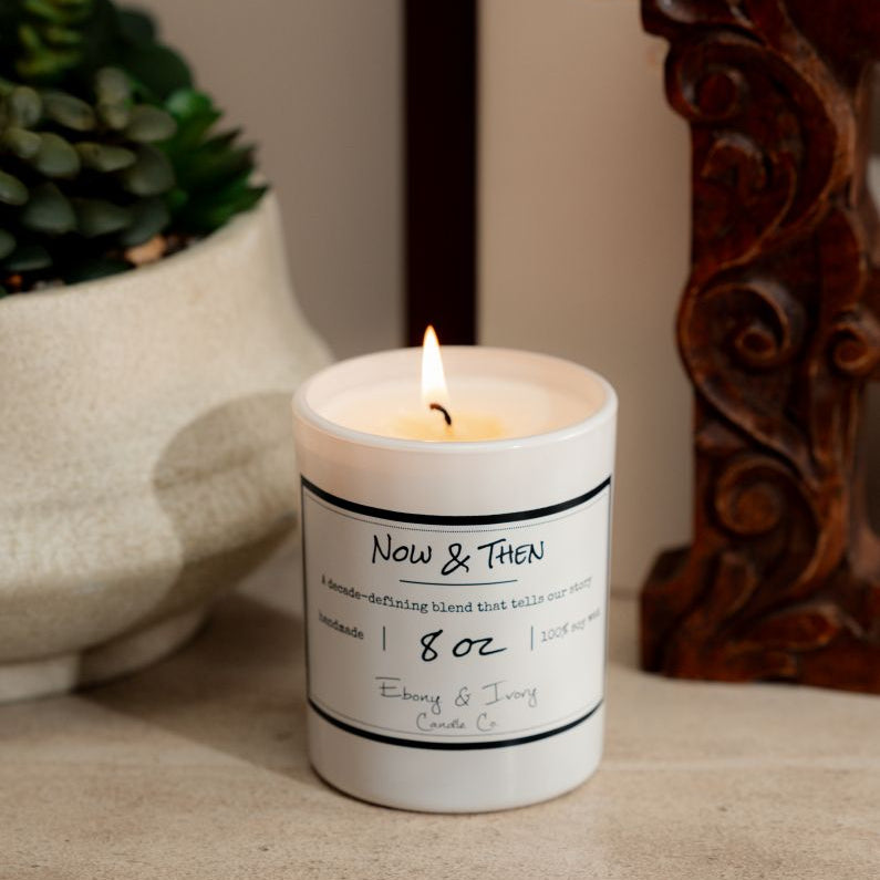 a white 8oz soy candle with a cotton wick sitting on a neutral background with a plant, two arts books, and a decorative wooden 'E'. The candle is labelled Now & Then, made by ebony & ivory candle co.