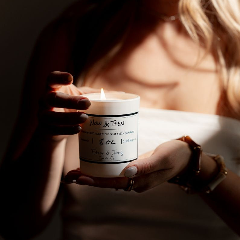 a white 8oz soy candle with a cotton wick, being held by a woman in a white dress with a spot light on it. The candle is labelled 'Now & Then' made by ebony & ivory candle co.