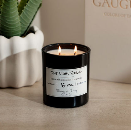 A black 16oz soy candle Labelled One Night Stand sitting on a table with a book and a potted plant. Made by Ebony & Ivory Candle Co in Lethbridge Alberta Canada
