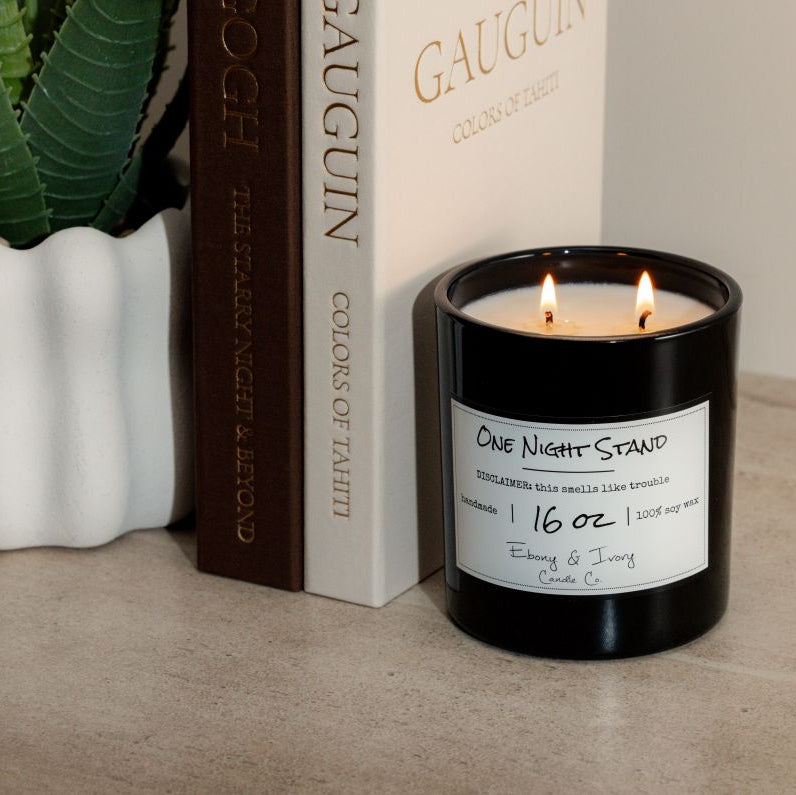 A black 16oz soy candle Labelled One Night Stand sitting on a table with a potted plant and two books. Made by Ebony & Ivory Candle Co in Lethbridge Alberta Canada