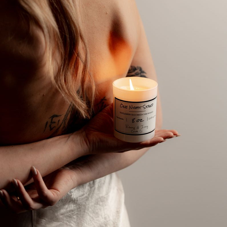 A white 8oz soy candle labelled One Night Stand, held by a woman behind her back. Made by Ebony & Ivory Candle Co in Lethbridge Alberta Canada