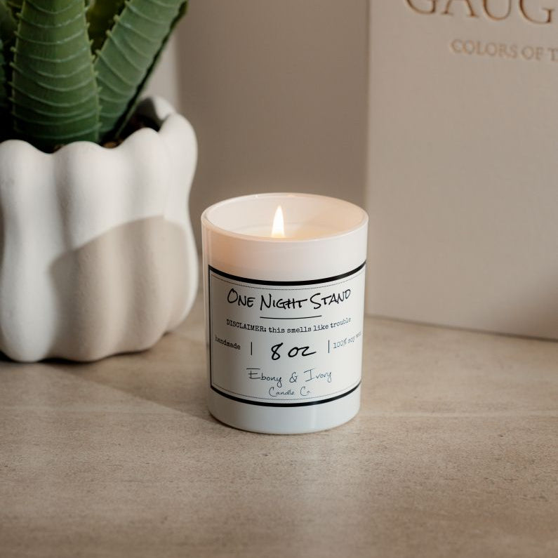 A white 8oz soy candle labelled One Night Stand, sittong a table with a potted plant and a book. Made by Ebony & Ivory Candle Co in Lethbridge Alberta Canada