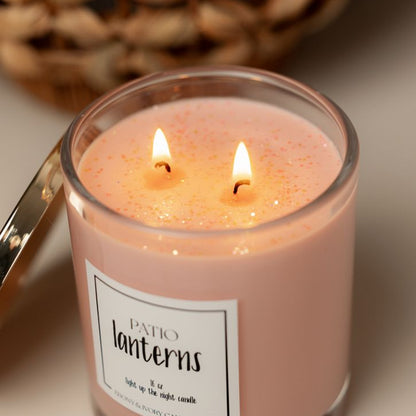 A clear 16oz soy candle with a gold lid labelled Patio Lanterns, sits on a table. The wicks are lit and there's soft peach coloured glitter on top. Made in Lethbridge Alberta