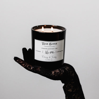 Candle labeled 'Rich Bitch' held by a hand in black lace glove against a gray background. Canadas soy candle non toxic.