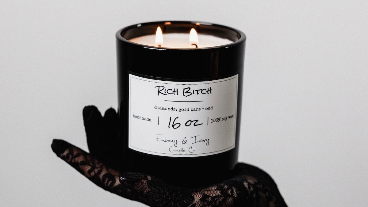 Candle labeled 'Rich Bitch' held by a hand in a black lace glove against a gray background