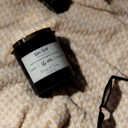 A black 16oz soy candle with a gold lid, labelled Rib Job, laying on a beach towel. Made in Lethbridge, alberta, canada