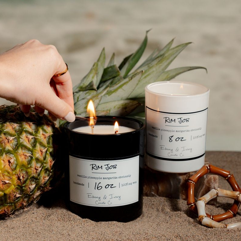 A white 8oz and a black 16oz soy wax candles labelled Rim Job, being lit on the beach, a pineapple beside them. Made in Lethbridge Alberta Canada