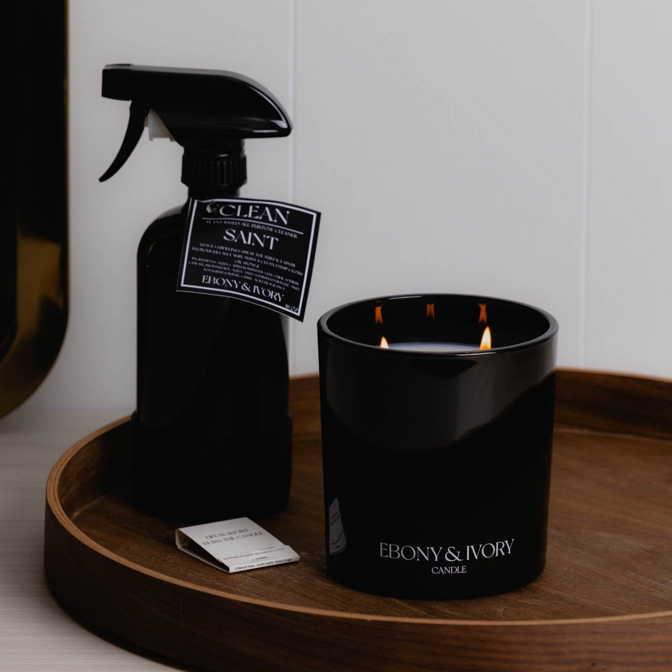 A black jar 18oz coconut soy candle with and a black 16oz spray bottle of all purpose cleaner with a black label named Saint, on a wooden tray with a clean white wall behind it, made by Ebony & Ivory Candle Co in Lethbridge, Alberta, Canada
