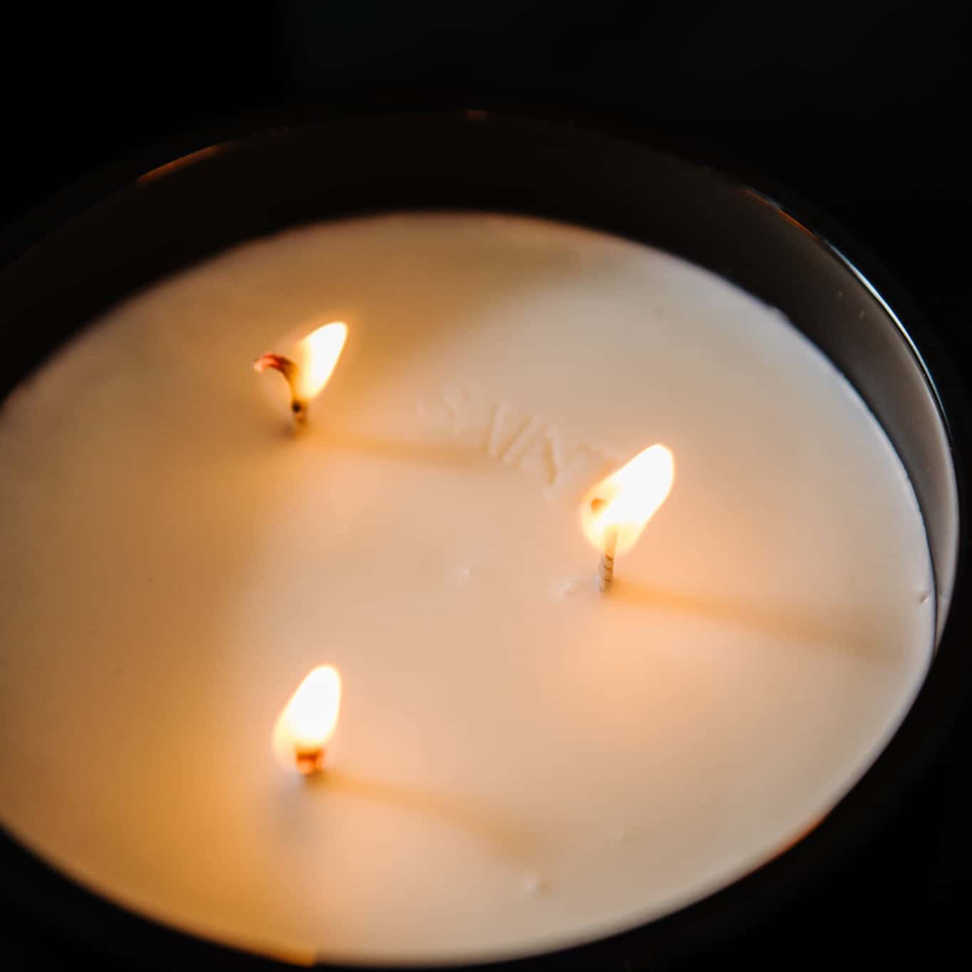 A top down view of a black jar 18oz coconut soy blend candle with the name "Sainy" imprinted on the wax inbetween three lit cotton wicks, on a black background, made by Ebony & Ivory Candle Co in Lethbridge, Alberta, Canada