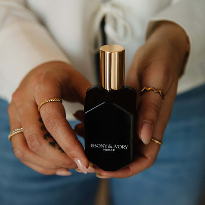 A black geometric perfume bottle with a gold lid and white text that says Ebony & Ivory Perfume, being held by someone with luxurious gold rings, made in lethbridge, alberta, canada 