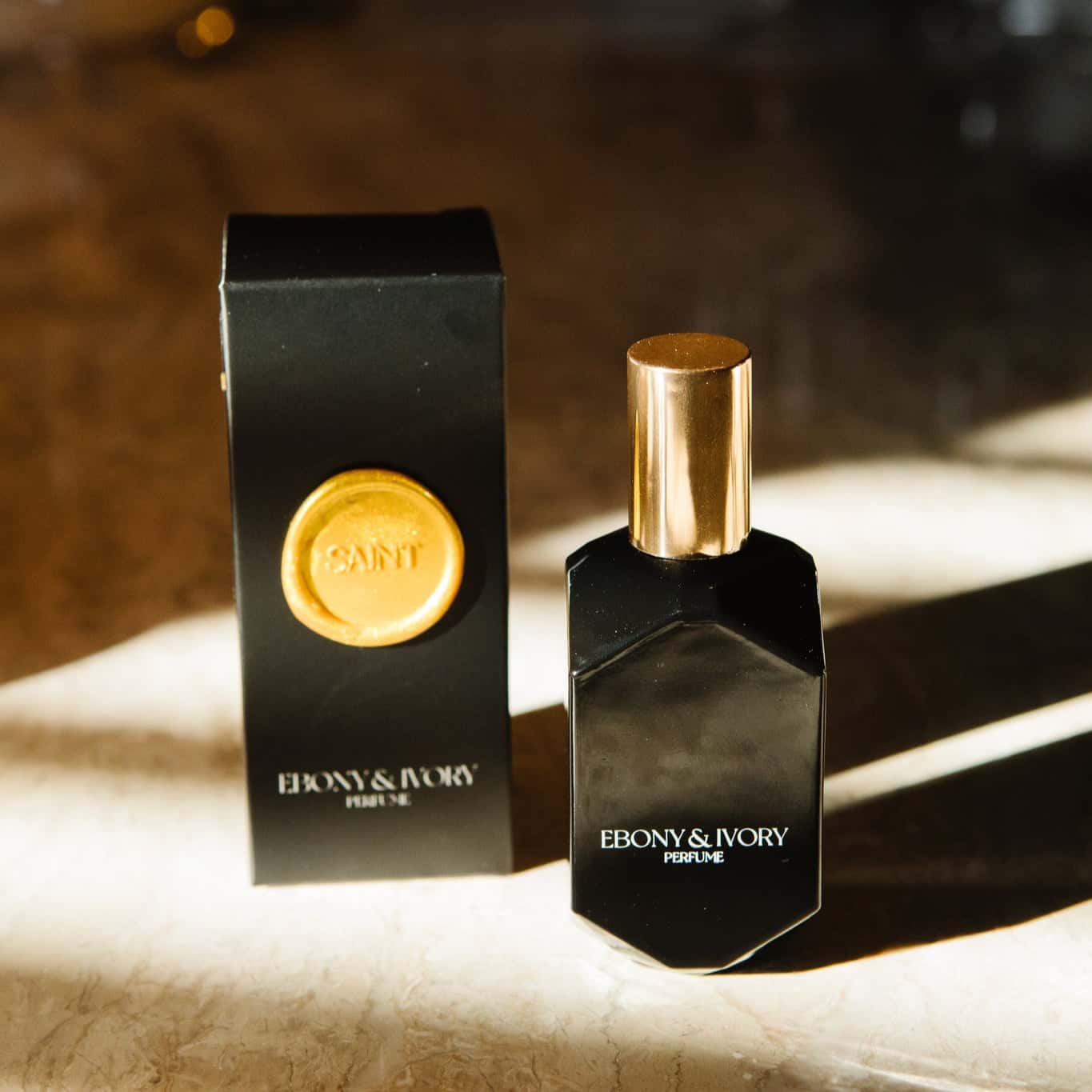 A black geometric perfume bottle with a gold lid and white text that says Ebony & Ivory Perfume, sitting on a granite counter top next to it's black box with a gold seal named SAINT, made in lethbridge, alberta, canada