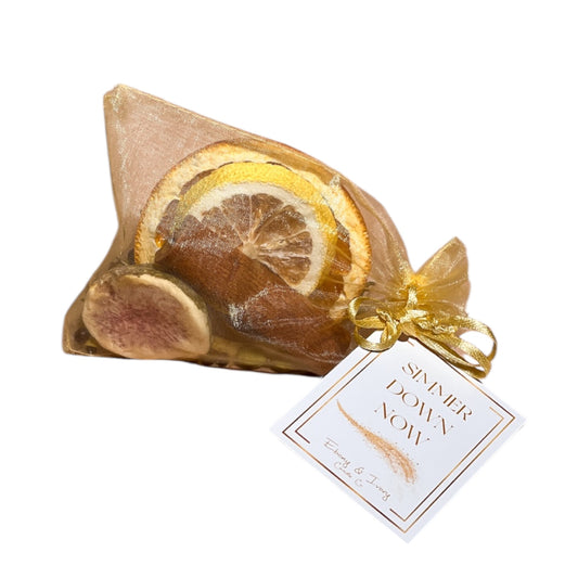 A gold organza bag filled with a dried lemon slice, orange slice, and a fig slice, with cinnamon, cardamom pods, rosemary, and peppercorns, tied up with a white label named Simmer Down Now, made by Ebony & Ivory Candle Co in Lethbridge, Alberta, Canada