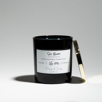 A black jar 16oz soy candle with a gold lid and a white labels named Ski Bum, on a white background, with blue coloured wax and silver glitter on top, made by Ebony & Ivory Candle Co in Lethbridge, Alberta, Canada.