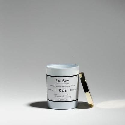 A white jar 8oz soy candle with a gold lid and a white labels named Ski Bum, on a white background, with blue coloured wax and silver glitter on top, made by Ebony & Ivory Candle Co in Lethbridge, Alberta, Canada.