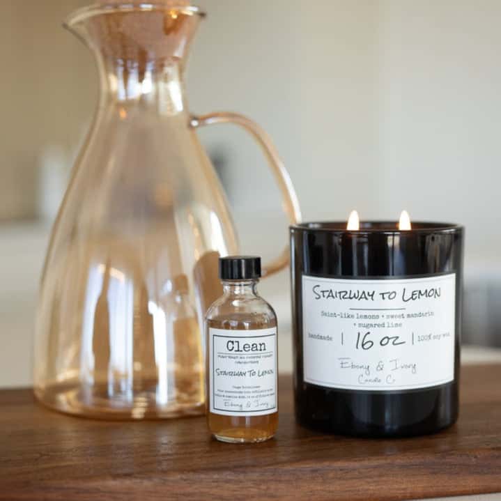 A black jar 16oz soy candle with a white label named Stairway to Lemon, on a wooden table next to an amber decanter, and a clear 2oz bottle of all purpose cleaner concentrate with a black cap and white label named Stairway to Lemon, made by Ebony & Ivory Candle Co in Lethbridge, Alberta, Canada