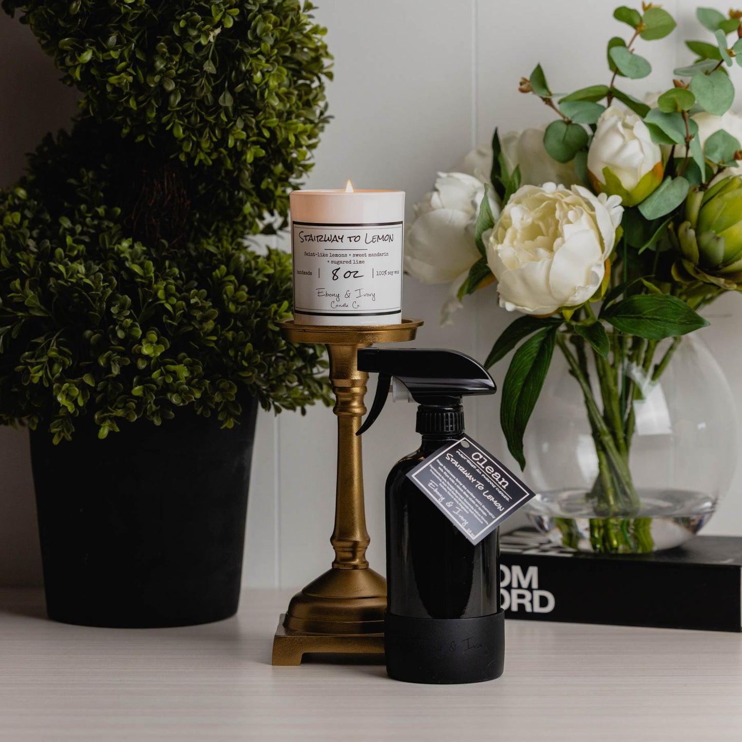 A white 8oz jar soy candle with a white label named Stairway to Lemon, on top of a golden candle holder, next to a black 16oz spray bottle of all purpose cleaner with a black rubber bottom and a black label named Stairway to Lemon, on a shelf. There's a potted shrub and a vase of flowers on top of a luxury brand book. Made by Ebony & Ivory Candle Co in Lethbridge, Alberta, Canada