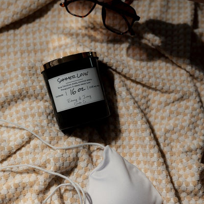 A black 16oz soy candle with a gold lid, labelled Summer Lovin, laying on a beach towel with a white bikini and sunglasses. Made in lethbridge alberta canada