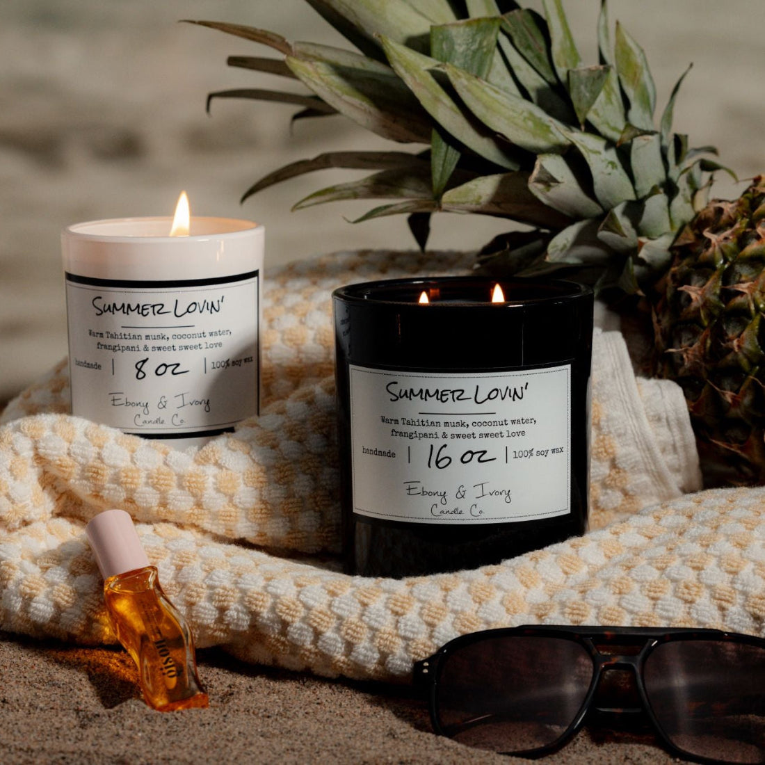 a white 8oz and a black 16oz soy candles labelled Summer Lovin, sitting on a towel on the beach with sunglasses and a pineapple.