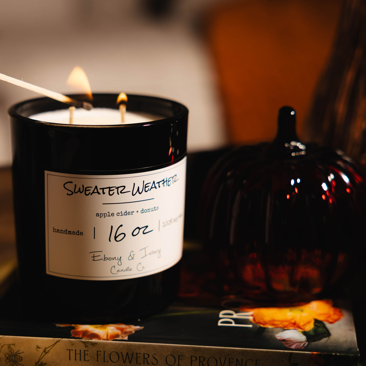 A black jar 16oz soy candle with a white label named Sweater Weather, on a stack of books with a glass pumpkin beside it. The two cotton wicks are being lit by a match stick. made by Ebony & Ivory Candle Co in Lethbridge, Alberta, Canada