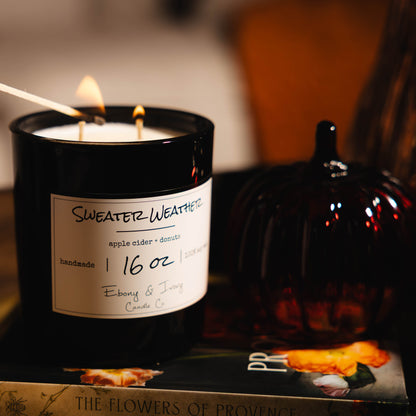 A black jar 16oz soy candle with a white label named Sweater Weather, on a stack of books with a glass pumpkin beside it. The two cotton wicks are being lit by a match stick. made by Ebony & Ivory Candle Co in Lethbridge, Alberta, Canada