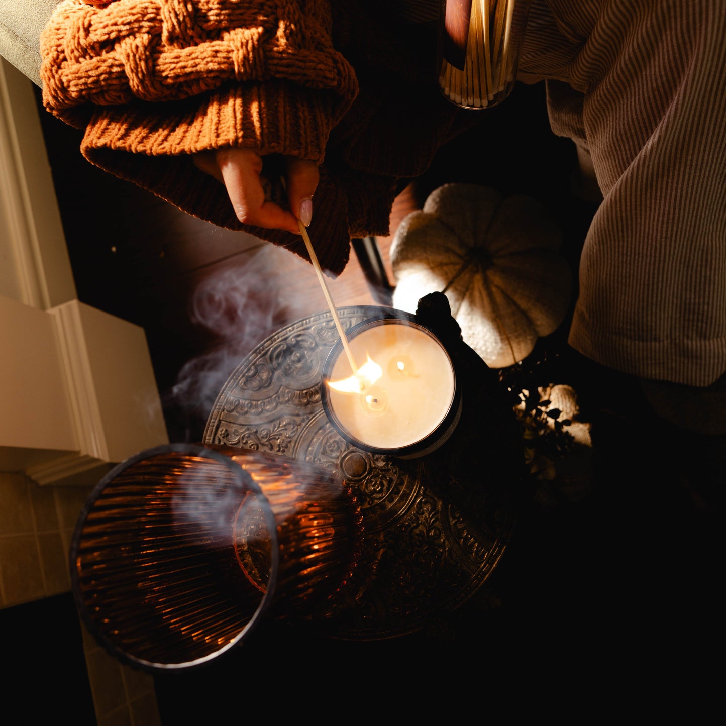 A top down view of a black jar 16oz soy candle with a white label named Sweater Weather, on a metal side table. The two cotton wicks are being lit by someone in a cozy sweater with a match stick. made by Ebony & Ivory Candle Co in Lethbridge, Alberta, Canada