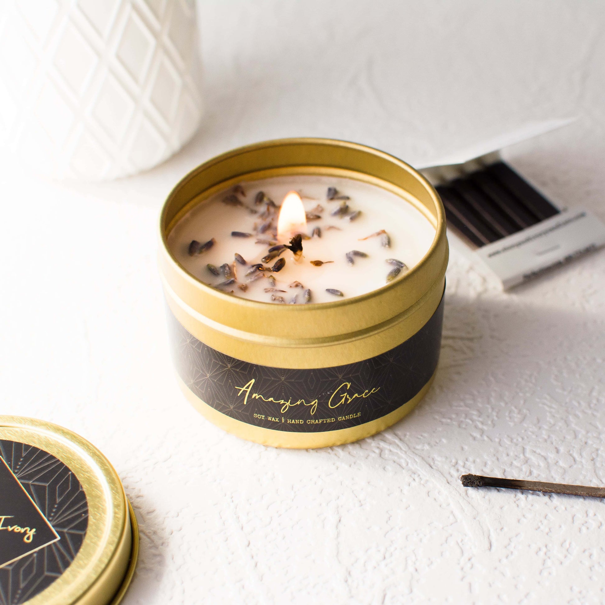 A 4oz gold tin soy candle with a black label that's named Amazing Grace with dried lavender buds on top, sits on a clean white table with a book of matches, made by Ebony & Ivory Candle Co in Lethbridge, Alberta, Canada