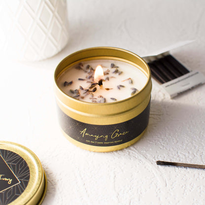 A 4oz gold tin soy candle with a black label that's named Amazing Grace with dried lavender buds on top, sits on a clean white table with a book of matches, made by Ebony & Ivory Candle Co in Lethbridge, Alberta, Canada