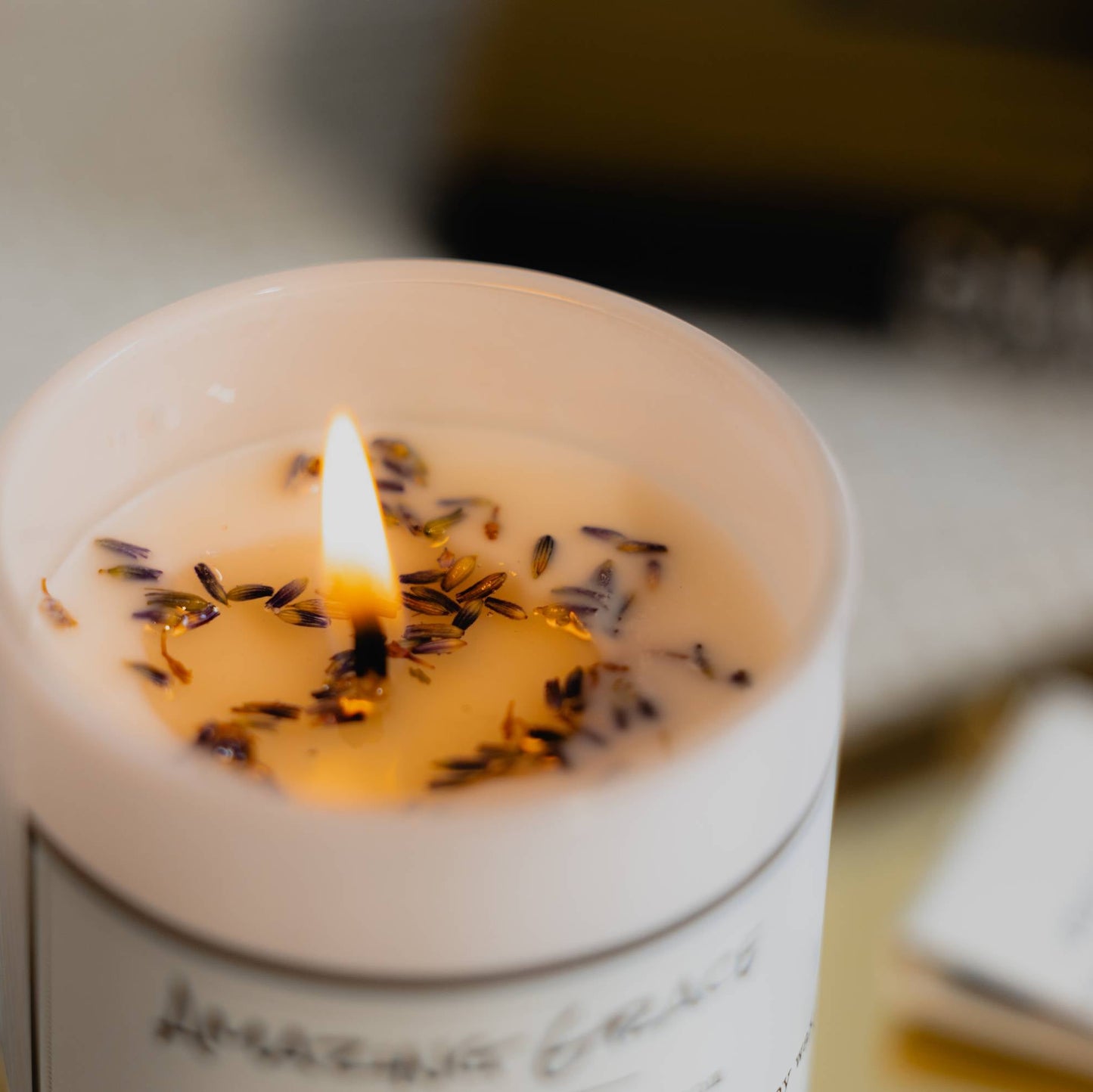 A top down shot of a white 8oz jar soy candle with a white label named Amazing Grace with dried lavender buds on top of the melted wax in a cozy, blurred indoor setting, made by Ebony & Ivory Candle Co in Lethbridge, Alberta, Canada
