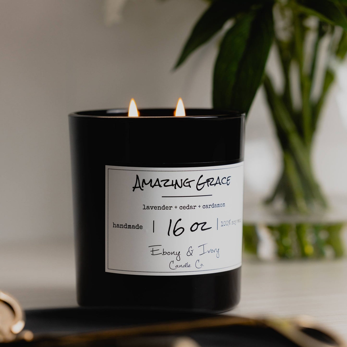 A black 16oz jar soy candle with a gold lid and a white label named Amazing Grace a white table with a clear vase of flowers and a black tray, made by Ebony & Ivory Candle Co in Lethbridge, Alberta, Canada