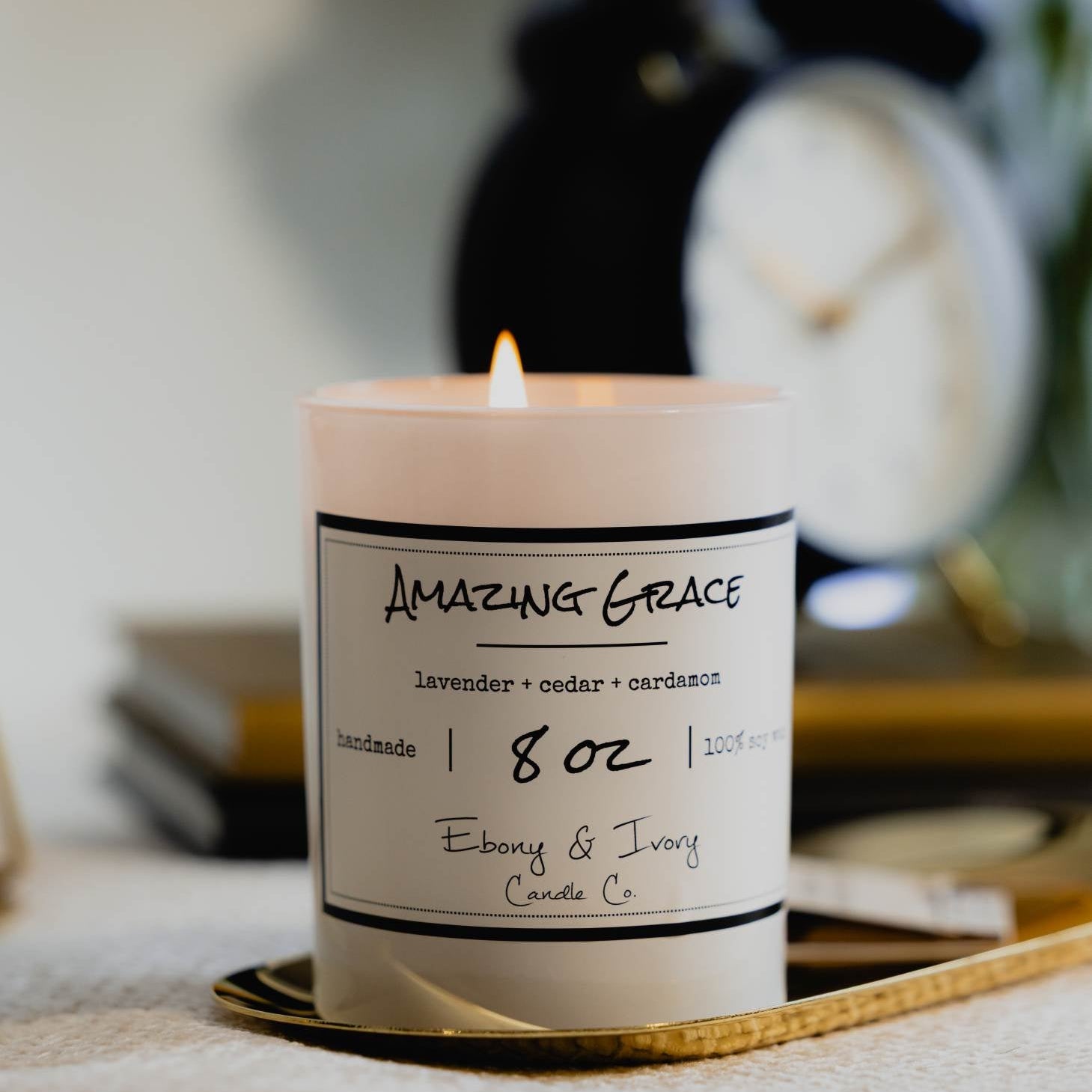 A white 8oz jar soy candle with a white label named Amazing Grace on a gold tray with a stack of books and a black alarm clock, made by Ebony & Ivory Candle Co in Lethbridge, Alberta, Canada