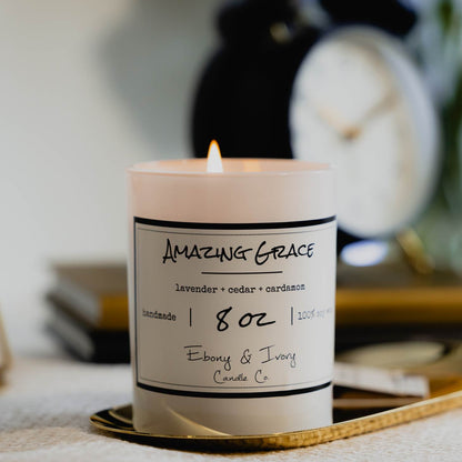 A white 8oz jar soy candle with a white label named Amazing Grace on a gold tray with a stack of books and a black alarm clock, made by Ebony & Ivory Candle Co in Lethbridge, Alberta, Canada