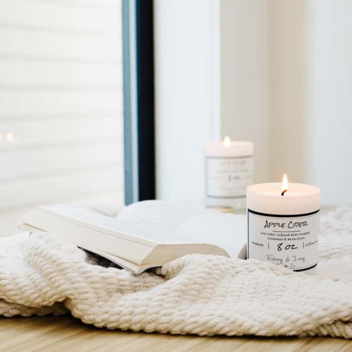 A white 8oz jar soy candle with a white label named Apple Cider in a windowsill with a cozy cream blanket and an open book next to it, made by Ebony & Ivory Candle Co in Lethbridge, Alberta, Canada
