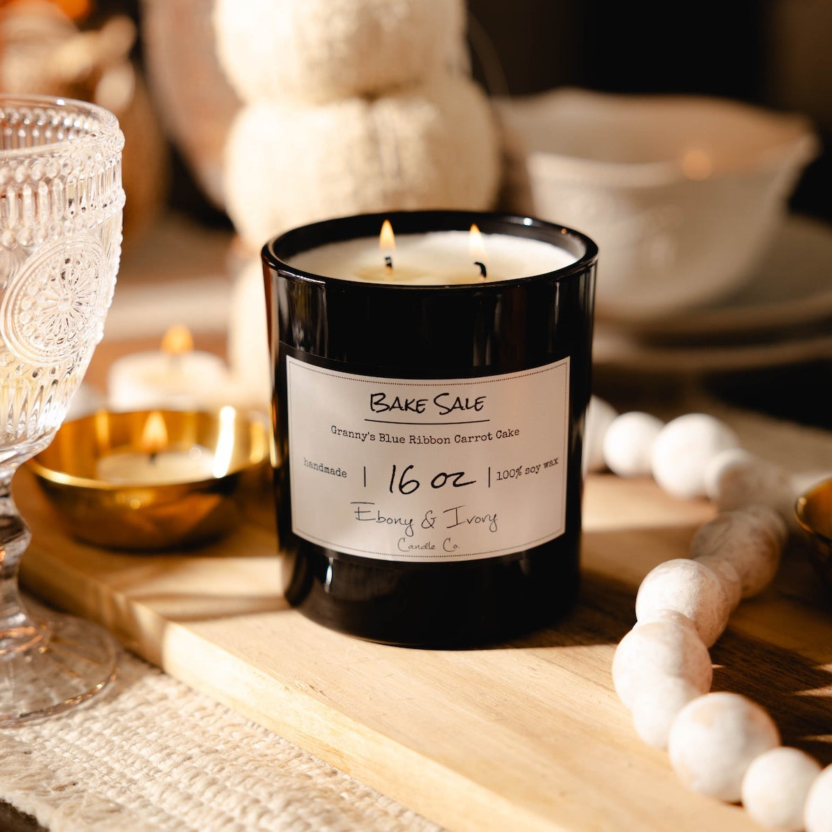Candle labeled 'Bake Sale' on a table with a glass, bottle, and decorative items.