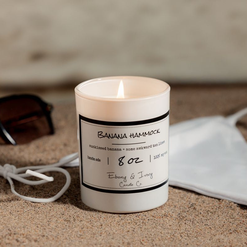 A white 8oz soy candle labelled Banana Hammock sitting on a beach. Made in Lethbridge, Alberta, Canada