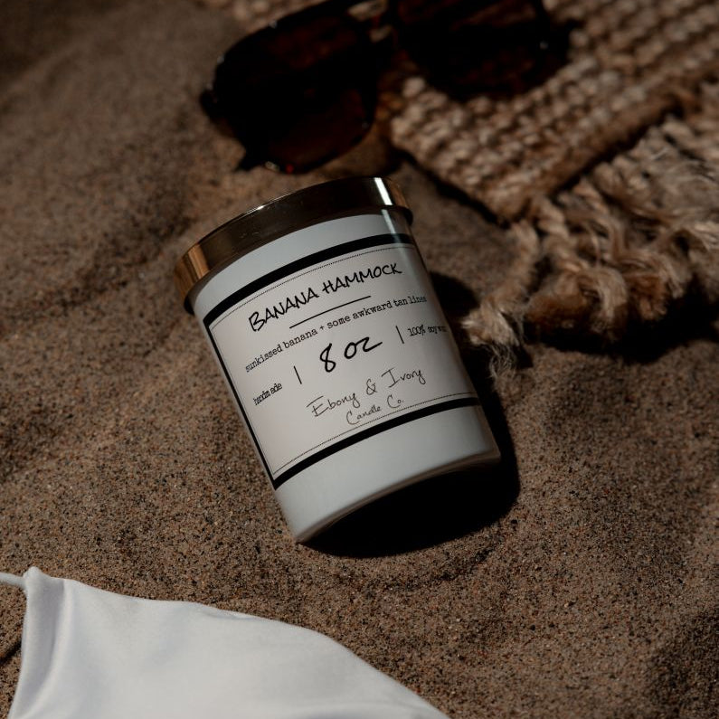 A white 8oz soy candle with a gold lid, labelled Banana Hammock, laying on the beach. Made in Lethbridge Alberta Canada