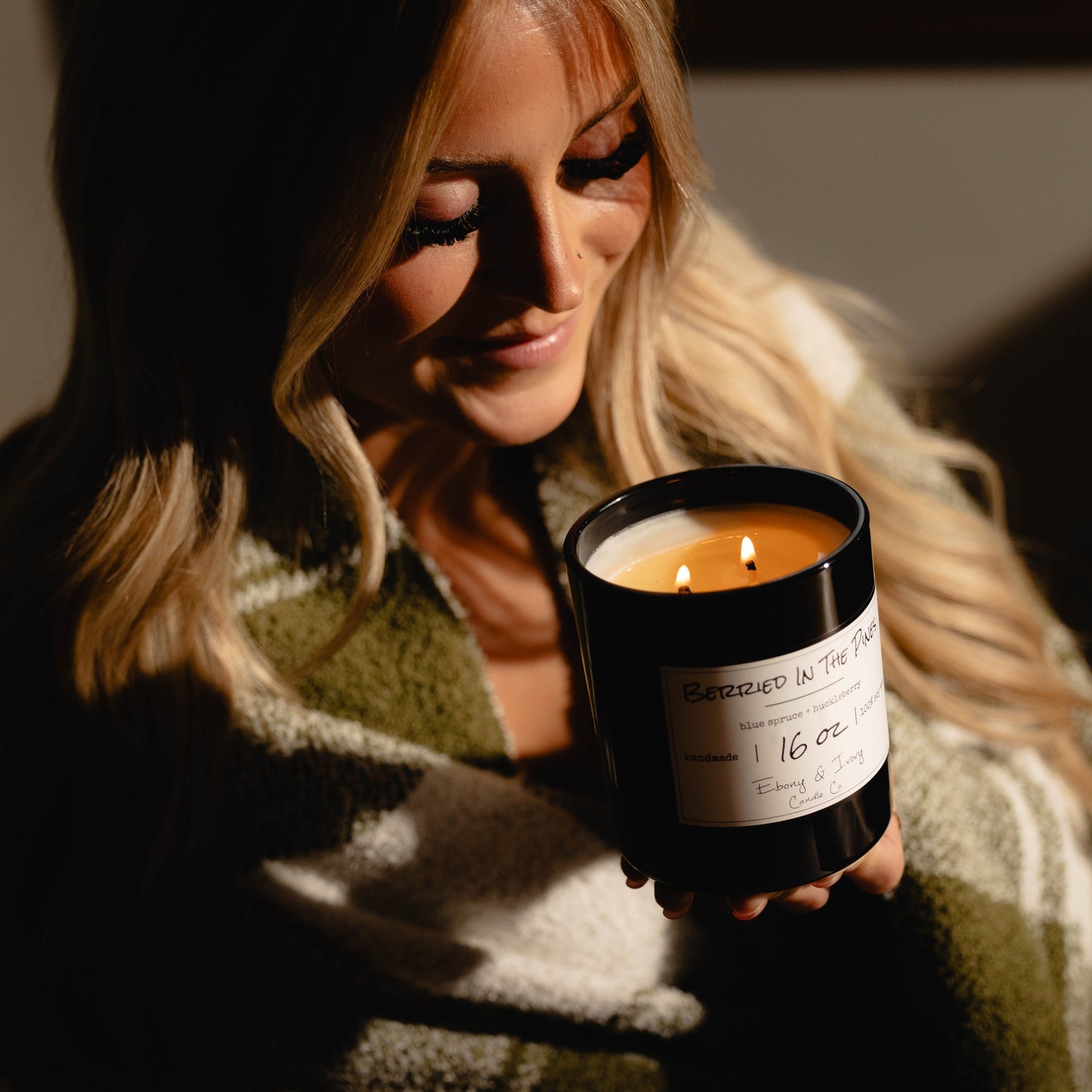 Woman holding a black 16oz soy candle with a gold lid and a white label, named 'Berried in the Pines' surrounded by warm lighting.