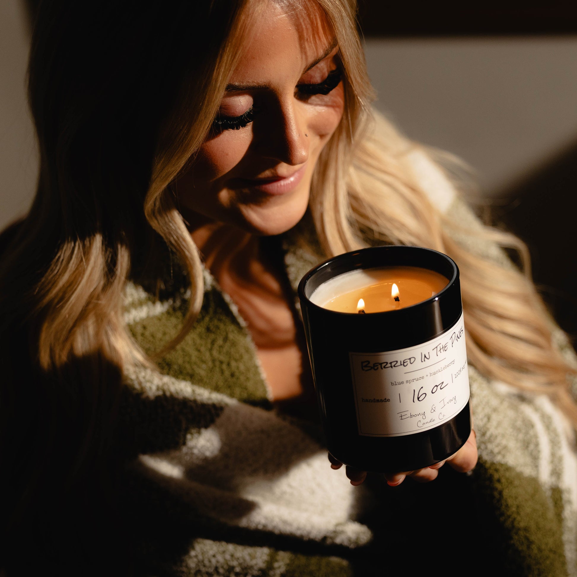 Woman holding a black 16oz soy candle with a gold lid and a white label, named 'Berried in the Pines' surrounded by warm lighting.