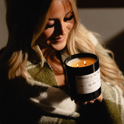 Woman holding a black 16oz soy candle with a gold lid and a white label, named 'Berried in the Pines' surrounded by warm lighting.