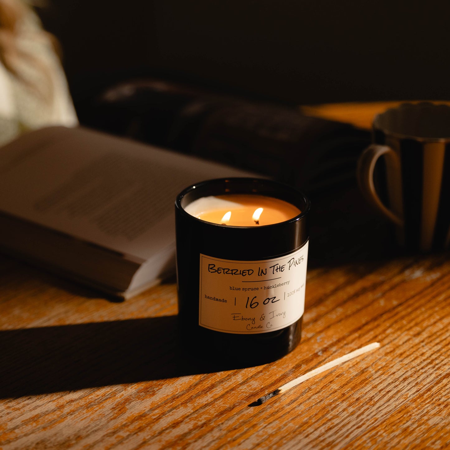 A black 16oz soy Candle with a gold lid and a white label named 'Berried in the Pines' on a wooden surface next to a book and mug