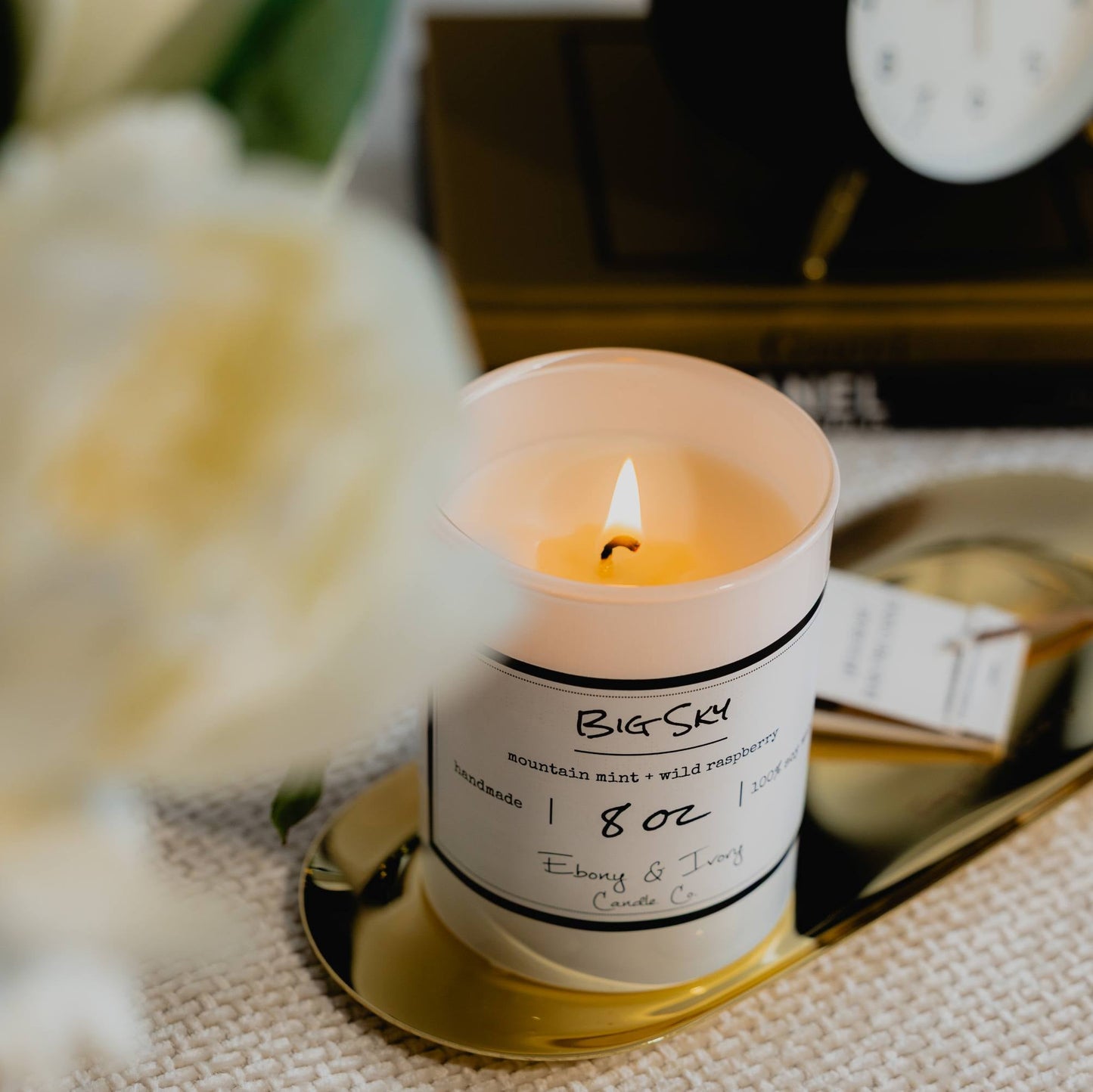 A white 8oz jar soy candle with a gold lid and a white label named Berried in the Pines, on a gold tray with books and a black alarm clock in the background, and white flowers in the foreground, made by Ebony & Ivory Candle Co in Lethbridge, Alberta, Canada