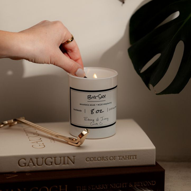 A white 8oz soy candle labelled Big Sky sitting on a stack of books with a pair of golden wick trimmers and a hand lighting the wick. Made in Lethbridge Alberta Canada