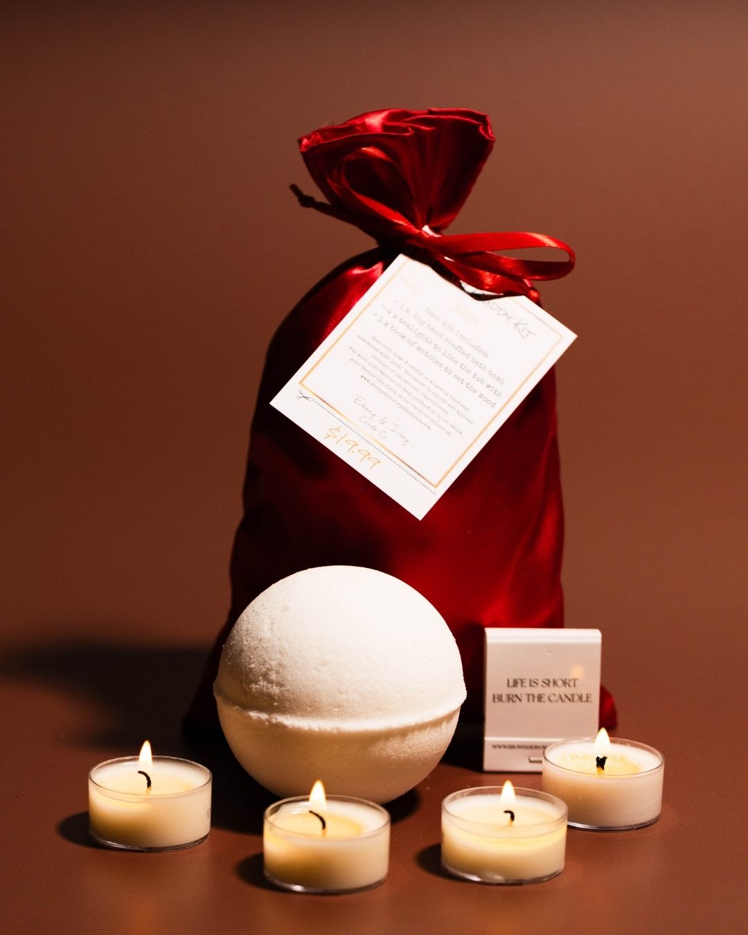 A red satin bag with a white hang tag sits behind a large bath bomb, a book of matches, and 4 tea lights.