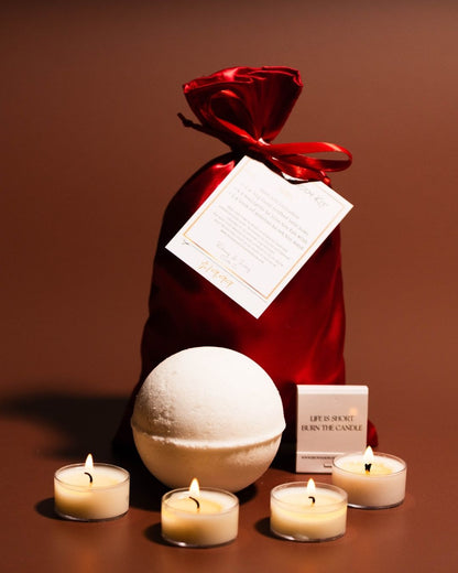 A red satin bag with a white hang tag sits behind a large bath bomb, a book of matches, and 4 tea lights.