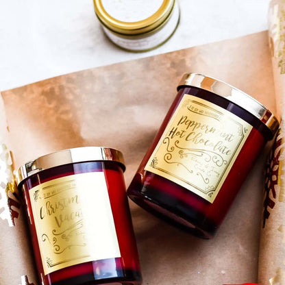 two red 8oz soy christmas candles with gold lids and old labels named "christmas vacation" and "peppermint hot chocolate" laying on christmas wrapping paper